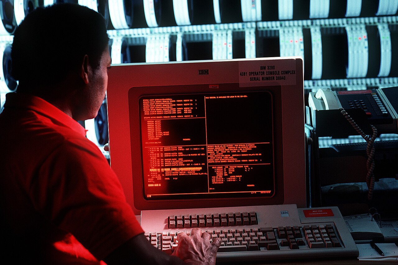 An operator at an IBM 4381 mainframe console, tape drives behind him